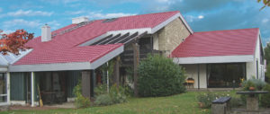 Modernes Haus mit rotem Dach, großen Fenstern und Steinwand, umgeben von Grünpflanzen und Rasen unter blauem Himmel.