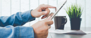 Hände bedienen ein Tablet, im Hintergrund stehen eine Kaffeetasse und einige Dokumente auf einem Tisch.