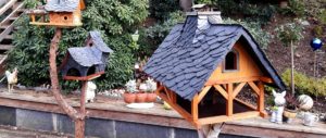Zwei dekorative Vogelhäuser mit dunklen Dächern stehen im Garten, umgeben von Pflanzen und Blumentöpfen.
