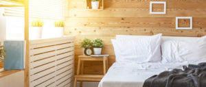 Helles Schlafzimmer mit Holzwänden, weißem Bett, Nachttisch, Pflanzen und Wecker, Sonnenlicht fällt durch das Fenster.