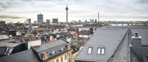 Blick über Dächer einer Stadt mit Fernsehturm und modernen Gebäuden im Hintergrund an einem bewölkten Tag.