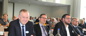 Drei Männer in Anzügen sitzen an einem Tisch in einem Konferenzraum, im Hintergrund sind weitere Teilnehmer zu sehen.