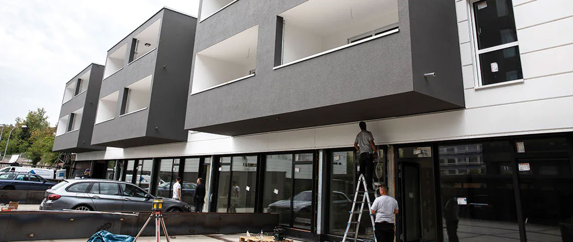 Moderne Wohnhausfassade mit großen Fenstern und Balkonen, graue Außenwände, spiegelnde Glasfront im Erdgeschoss, teilweise sichtbares geparktes Auto davor.