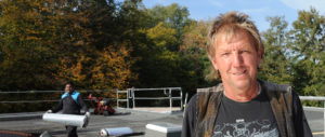Dachlive_Rund-um-den-Betrieb_Personal_Juergen-Teuteberg_Hero_neu Ein Bauarbeiter steht auf einer Baustelle im Freien, umgeben von Bäumen und Arbeitsgeräten, mit sonnigem Wetter im Hintergrund.
