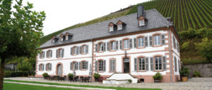 Ein großes, weißes Gebäude mit grauem Dach und vielen Fenstern, davor Tische und Stühle, im Hintergrund ein begrünter Hang.