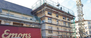 Ein mehrstöckiges Gebäude mit Baugerüst, Arbeiter auf dem Gerüst, Fassade wird renoviert oder saniert, unten ein rotes Fahrzeug abgebildet.