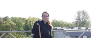 Eine Frau mit Brille und schwarzer Jacke steht draußen, hält einen Holzstock, im Hintergrund Bäume und Gebäude bei Tageslicht.