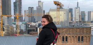 Eine Person steht auf einem Dach und lächelt, mit einer Stadtansicht und Baukränen im Hintergrund.