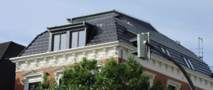 Modernes Dachgeschoss mit großen Fenstern und Solarpaneelen, auf einem historischen Gebäude mit dekorativen Fassadenelementen, teilweise von einem Baum verdeckt.