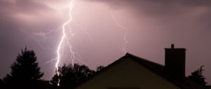 Blitze am Nachthimmel über einem dunklen Hausdach und Bäumen, der Himmel ist hell erleuchtet.