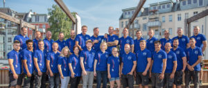 Eine Gruppe von Menschen in blauen Poloshirts steht draußen auf einer Brücke und posiert gemeinsam für ein Gruppenfoto bei sonnigem Wetter.