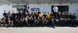 Eine Gruppe von Menschen in Arbeitskleidung sitzt und steht vor einem Gebäude mit Schildern, sie lächeln und wirken als Team.