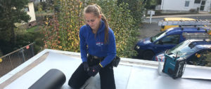 Dach-Live-Dachdeckerin-Justine-Schmid-Natalie-Boll-Hero Eine Frau mit Handschuhen kniet auf einem Dach und trägt eine blaue Arbeitsjacke, im Hintergrund sind Bäume und ein blaues Auto zu sehen.