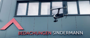 Dach-Live-Digitalisierung-Sindermann-Drohne-Hero Eine Drohne fliegt vor einem modernen Gebäude mit der Aufschrift „ÜBERWACHUNGEN SIND“, wobei „ÜBERWACHUNGEN“ in roten Buchstaben hervorgehoben ist.