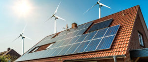 Solarmodule auf einem Hausdach, im Hintergrund mehrere Windräder vor blauem Himmel, Symbol für erneuerbare Energien und umweltfreundliche Stromerzeugung.