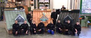 Eine Gruppe von acht Personen posiert in Arbeitskleidung in einer Werkstatt vor Modellen von Dachkonstruktionen aus Holz und Ziegel.