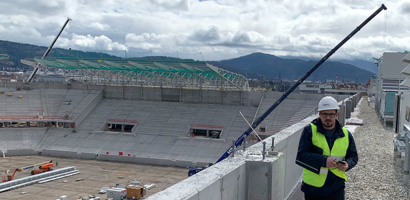 Blick auf eine Stadionbaustelle mit Tribünen, Baukran und Bergen im Hintergrund unter bewölktem Himmel.