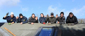 Fünf Menschen winken und lächeln auf einem Dach bei klarem, blauem Himmel.