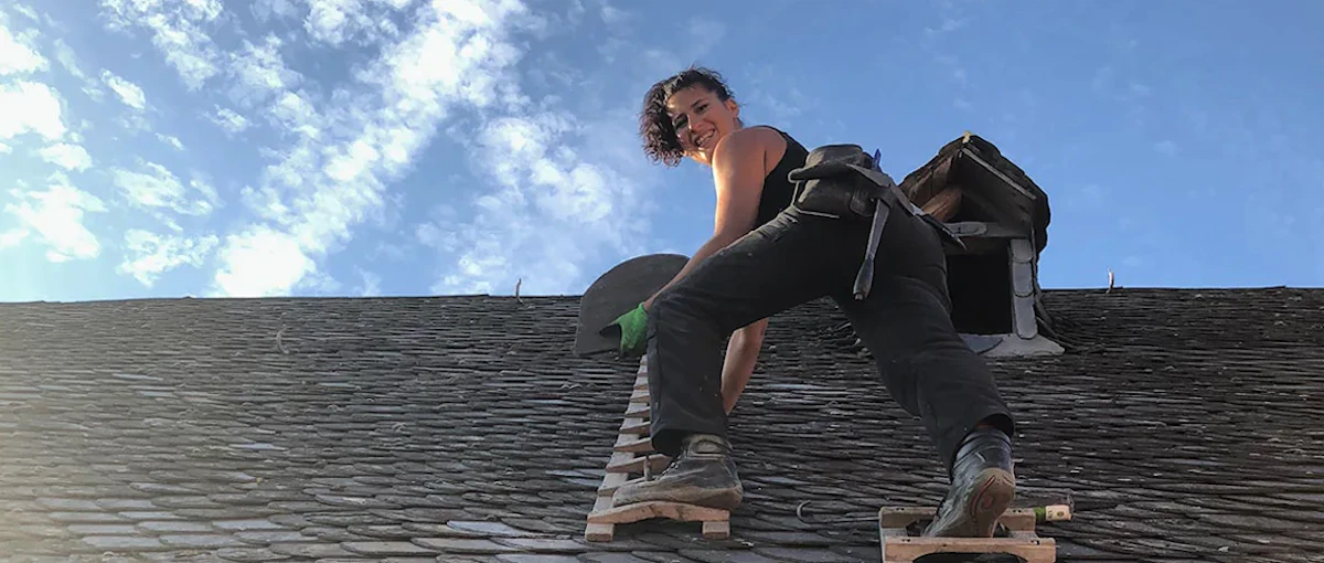 Eine Frau mit kurzem Haar und grünem Handschuh lächelt, während sie auf einer Dachleiter bei sonnigem Himmel arbeitet.