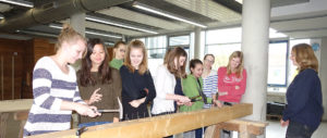Mehrere junge Frauen stehen an einem langen Holzbrett und schlagen Nägel ein, in einem hellen Raum mit großen Fenstern.