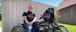 Dach-Live-Betriebsportrait-Dachdecker-Uwe-Horstmann-Hero Ein Mann mit Brille sitzt auf einem schwarzen Motorrad, trägt ein schwarzes Harley-Davidson-T-Shirt und blaue Jeans. Im Hintergrund eine Betonwand und blauer Himmel.
