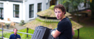 Junger Mann mit lockigem Haar hält Dachziegel, im Hintergrund Gebäude mit begrüntem Dach.