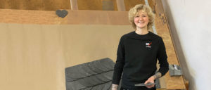 Eine lächelnde Person mit lockigem blondem Haar steht in einem Kletterraum vor einer Kletterwand mit Matten am Boden.