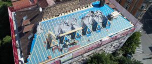 Luftaufnahme einer Baustelle auf einem Dach mit mehreren Arbeitern, Baugerüsten und abgedeckten Holzstrukturen.