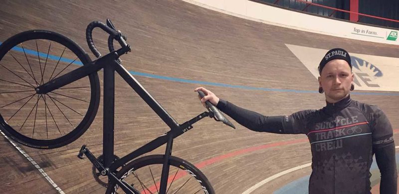 Dachdecker-Etienne-Sikora-Rennrad-Hero Person hält ein schwarzes Fahrrad auf einer Holzbahn im Velodrom, blaue und rote Linien sichtbar.