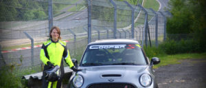 Eine Person in gelber Sicherheitskleidung steht neben einem silbernen Mini Cooper S auf einer Rennstrecke mit Sicherheitszaun im Hintergrund.