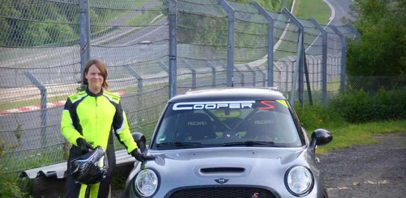 Dachdeckermeisterin-Coletta-Glitz-Mini-Cooper-Hero Eine Person in gelber Sicherheitskleidung steht neben einem silbernen Mini Cooper S auf einer Rennstrecke mit Sicherheitszaun im Hintergrund.