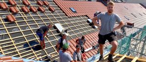 Mehrere Arbeiter decken ein Hausdach mit roten Ziegeln. Manche stehen, andere knien, während sie die Dachkonstruktion befestigen.