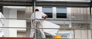 Ein Bauarbeiter bringt Dämmplatten an einer Hausfassade an. Ein Eimer steht neben ihm auf dem Gerüst.