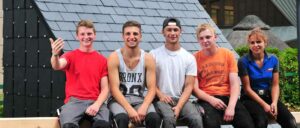 Zwei junge Männer sitzen nebeneinander vor einer Hauswand, einer trägt ein Tanktop, der andere ein weißes T-Shirt und eine Baseballkappe, beide lächeln.