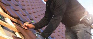 Eine Person arbeitet mit gebeugtem Arm auf einem Dach mit roten Dachziegeln und trägt dunkle Kleidung.