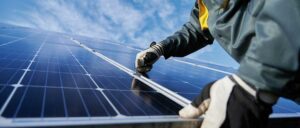 Eine Person mit Arbeitshandschuhen installiert ein Solarpanel unter blauem Himmel.