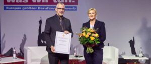 Zwei Personen bei einer Preisverleihung, eine hält eine Urkunde, die andere einen Blumenstrauß. Im Hintergrund ein Tisch mit Wasserflasche und Gläsern.