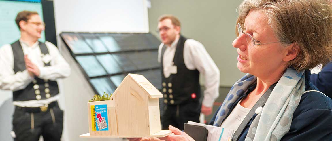 ZVDH-Pressesprecherin Claudia Büttner präsentiert am Messe-stand ein Miniatur-Holzhaus mit Gründach und PV-Anlage.