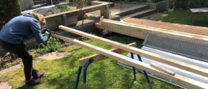 Holzbalken liegen auf einer Wiese, einige sind gestapelt, andere sind zu einer Struktur angeordnet. Im Hintergrund sind eine Betonmauer und Gartenwerkzeuge zu sehen.