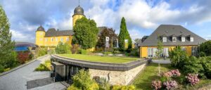 Ein gepflegter Garten mit Rasen, Bäumen und drei wehenden Fahnen vor einem großen gelben Gebäude, bewölkter Himmel im Hintergrund.