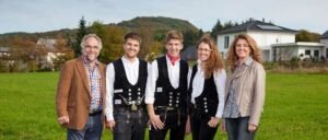 Ein junger Mann trägt traditionelle deutsche Kleidung mit rotem Halstuch und steht draußen vor einer hügeligen Landschaft.