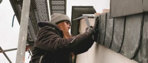 Eine Frau mit Mütze arbeitet draußen an einem Briefkasten oder Schaltkasten.
