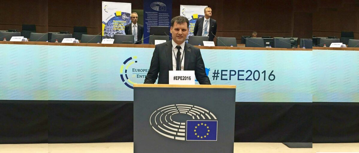eu-parlament-Helmut-Zimmer-Hero Ein Mann im Anzug spricht bei einer Veranstaltung mit Mikrofon, Hashtag #EPE201 und europäischem Parlamentslogo im Hintergrund.