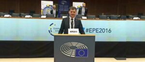 Ein Mann im Anzug spricht bei einer Veranstaltung mit Mikrofon, Hashtag #EPE201 und europäischem Parlamentslogo im Hintergrund.