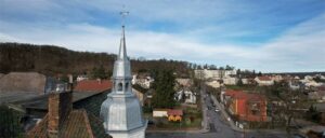 Dachdecker-Christian-Kallies-fertiger-Turm-Hero Kleinstadt mit Wohnhäusern, grünem Baum in der Mitte, Hügel im Hintergrund und teils bewölktem Himmel.