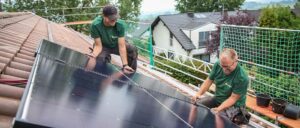 Person installiert Solarpaneele auf einem Hausdach, im Hintergrund weitere Häuser und Bäume.