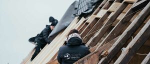 Eine Person mit einem dunklen Mantel und Schal steht draußen vor einem Holzgebäude mit Dachziegeln, Rückansicht, winterliche Stimmung.