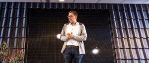 Ein Mann mit Brille, weißem Hemd und grauer Jacke steht und hält die Hände zusammen, Hintergrund mit dunklen vertikalen Linien.