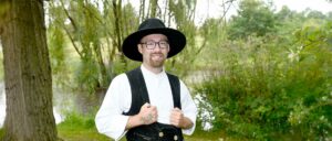 auf-der-Walz-Dachdeckergeselle-Luca-Umlauf Ein Mann mit Brille und schwarzem Hut trägt traditionelle Kleidung und steht draußen im Grünen.