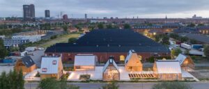 Moderne Gewächshäuser mit Glaswänden im Vordergrund, dahinter ein großes dunkles Gebäude und eine Stadtlandschaft im Hintergrund bei bewölktem Himmel.